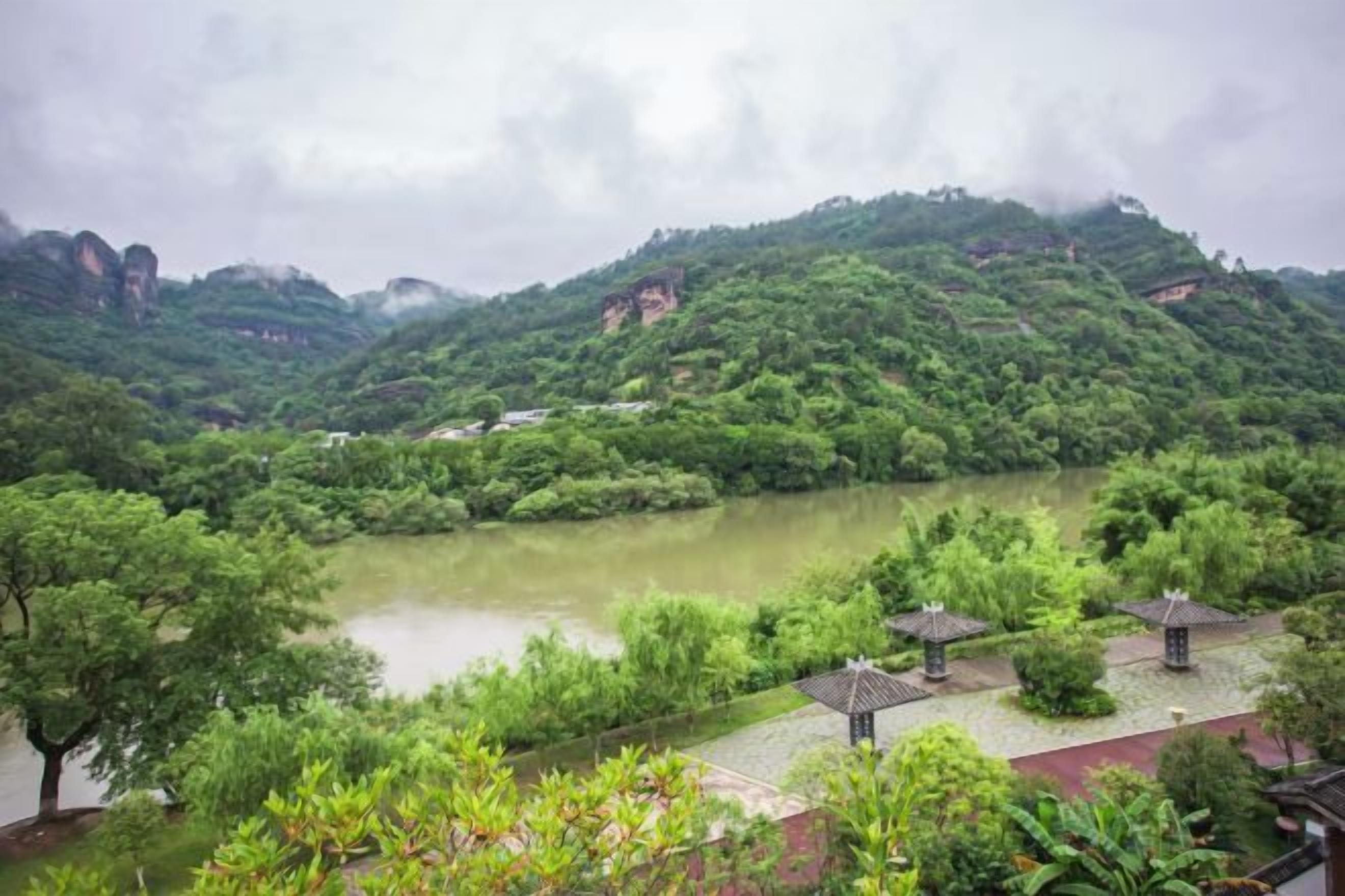 A river surrounded by lush greenery and hills, with a pavilion and a red-roofed building in the foreground.