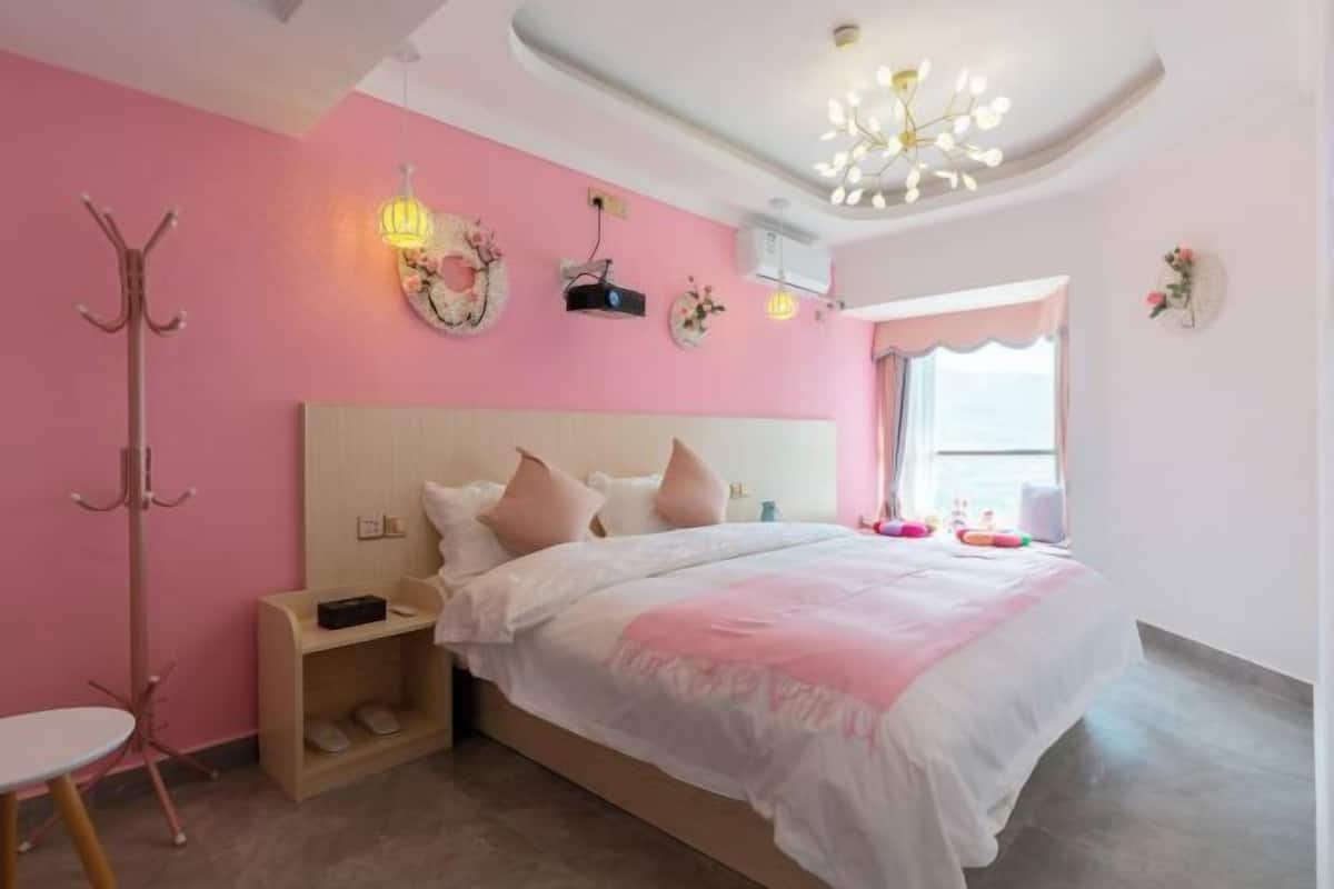 Ein Schlafzimmer mit rosa Wand, einem Holzkopfende, einem Bett mit weißer und pinkfarbener Bettwäsche, einem Nachttisch, einem Kleiderständer und einem Fenster mit Vorhängen.