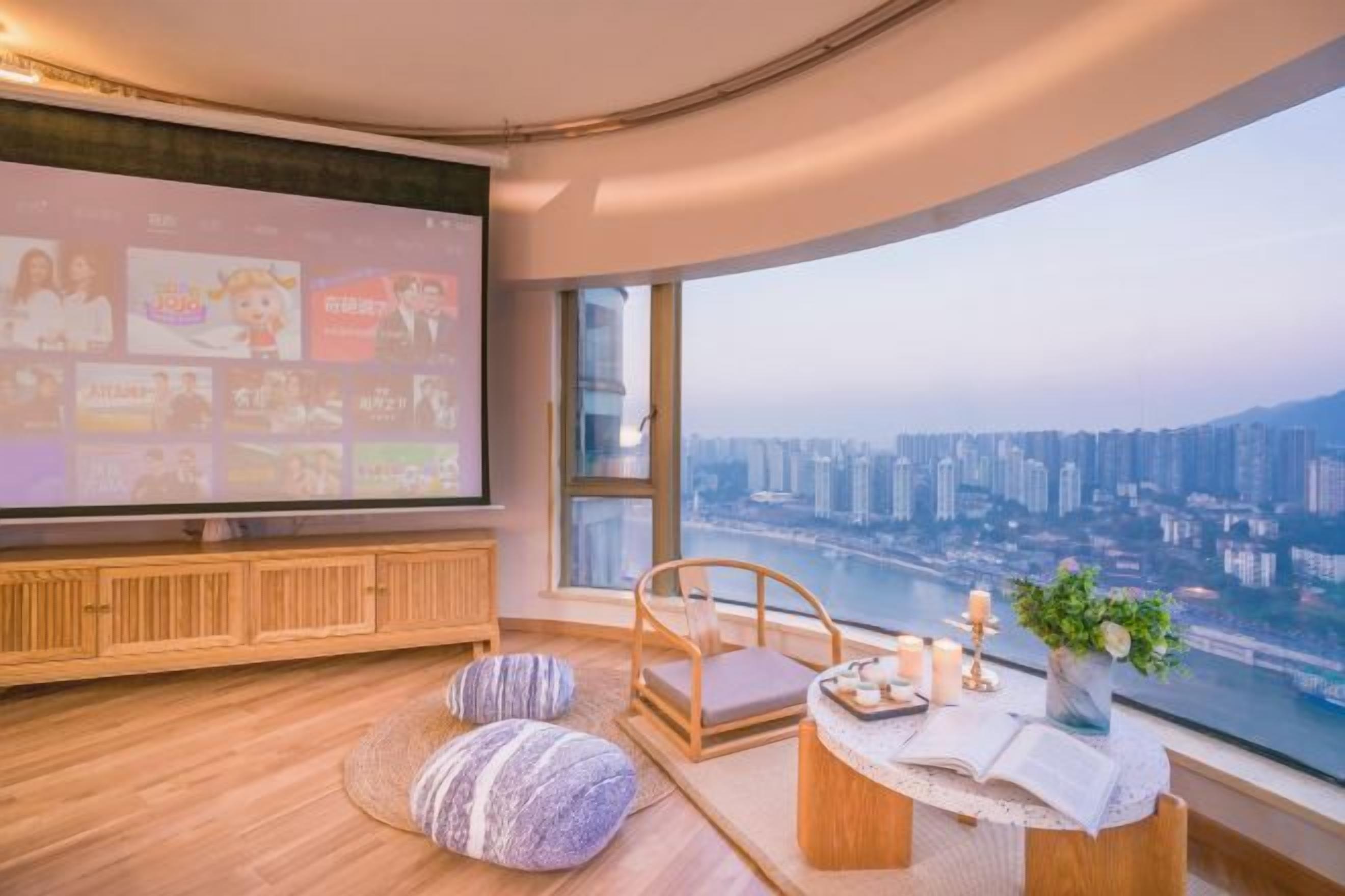 A modern living room with a large TV, comfortable seating, a small table with candles, and a view of a cityscape.