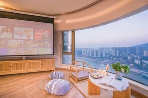 A modern living room with a large TV, comfortable seating, a small table with candles, and a view of a cityscape.