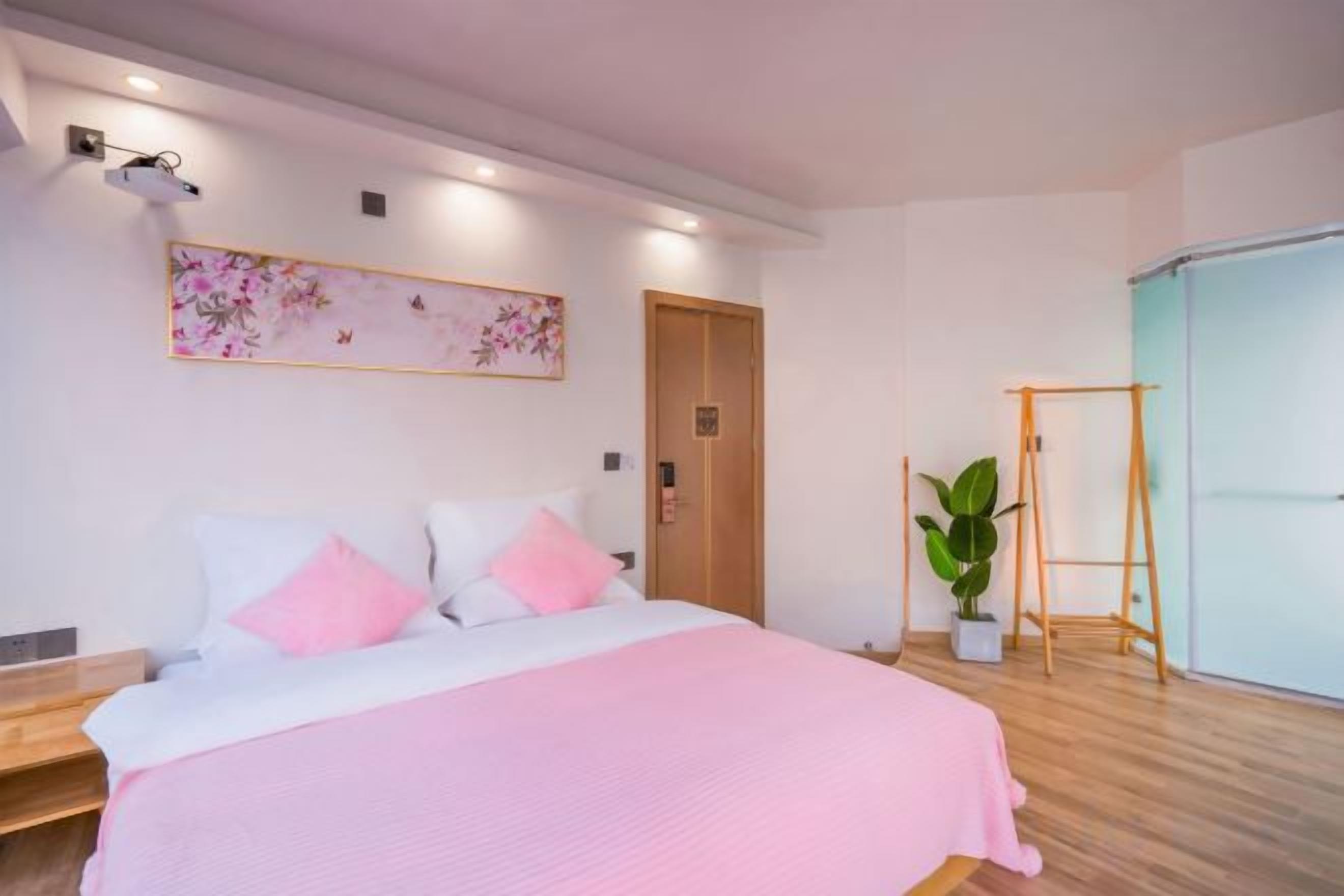 A bedroom with a bed, a wooden floor, a wall-mounted artwork, a door, and a plant.