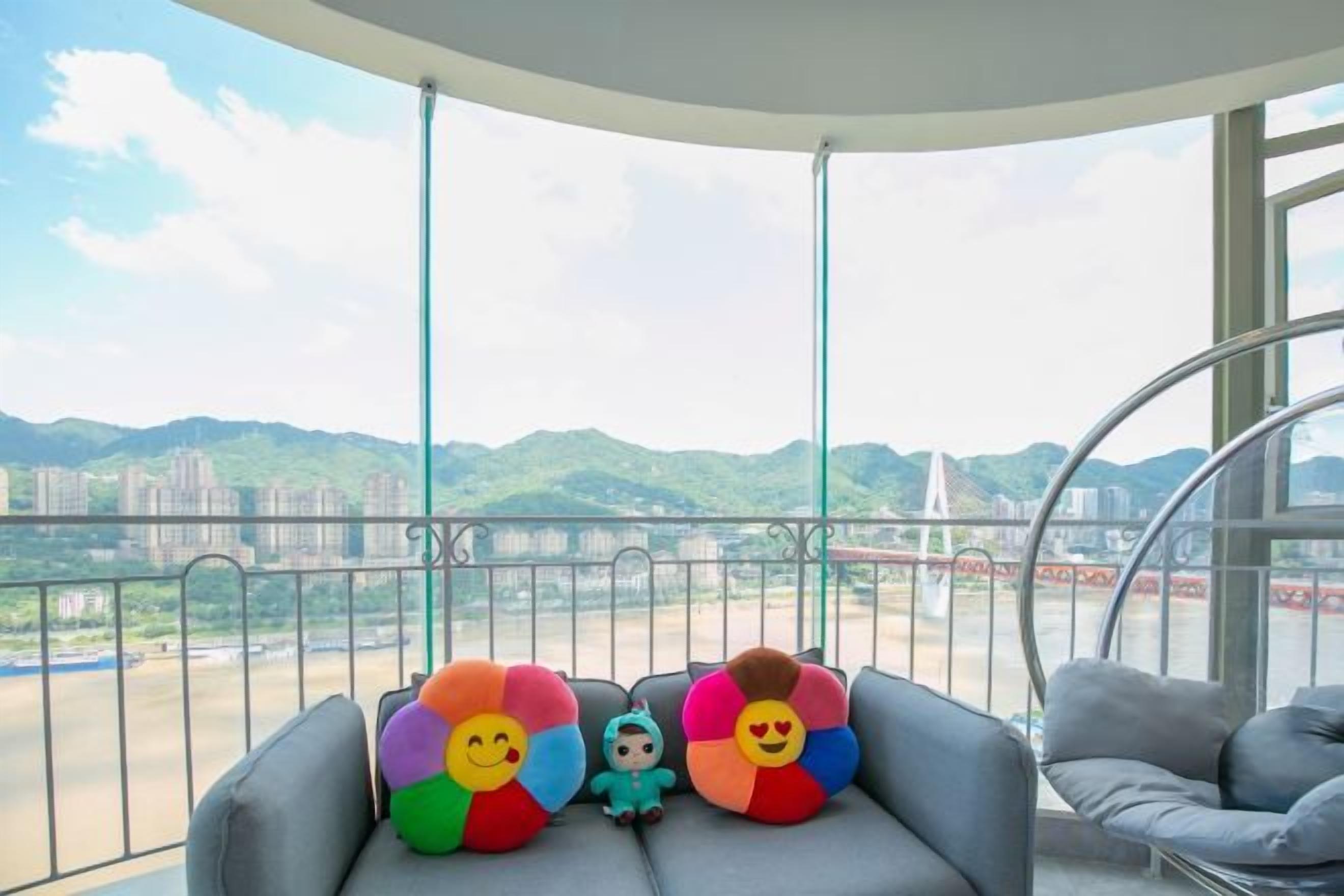 A balcony with a sofa and two colorful pillows, overlooking a cityscape with mountains and a bridge.