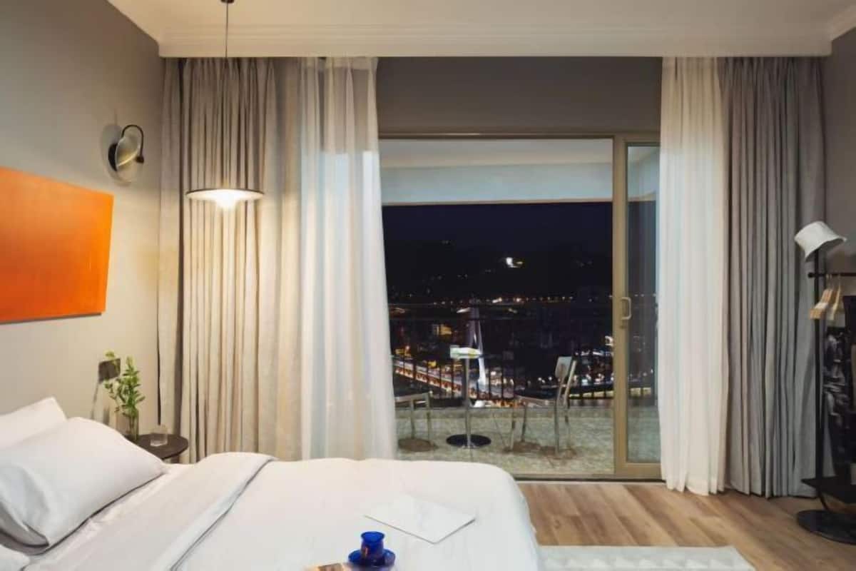 Ein Hotelzimmer mit Bett, Nachttisch, Lampe, einem Fenster mit Vorhängen und Blick auf nächtliche Stadtansichten.