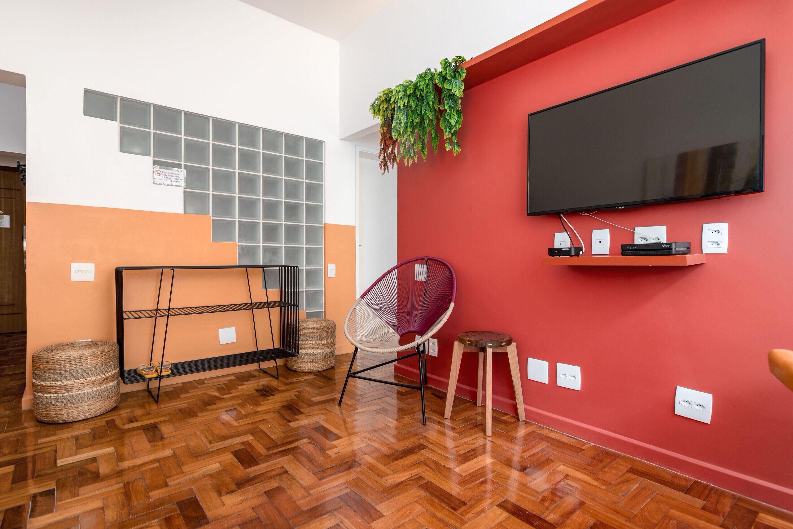 A modern living room with a herringbone wooden floor, a red accent wall, a mounted flat-screen TV, a wicker basket, a woven chair, and a small wooden stool.