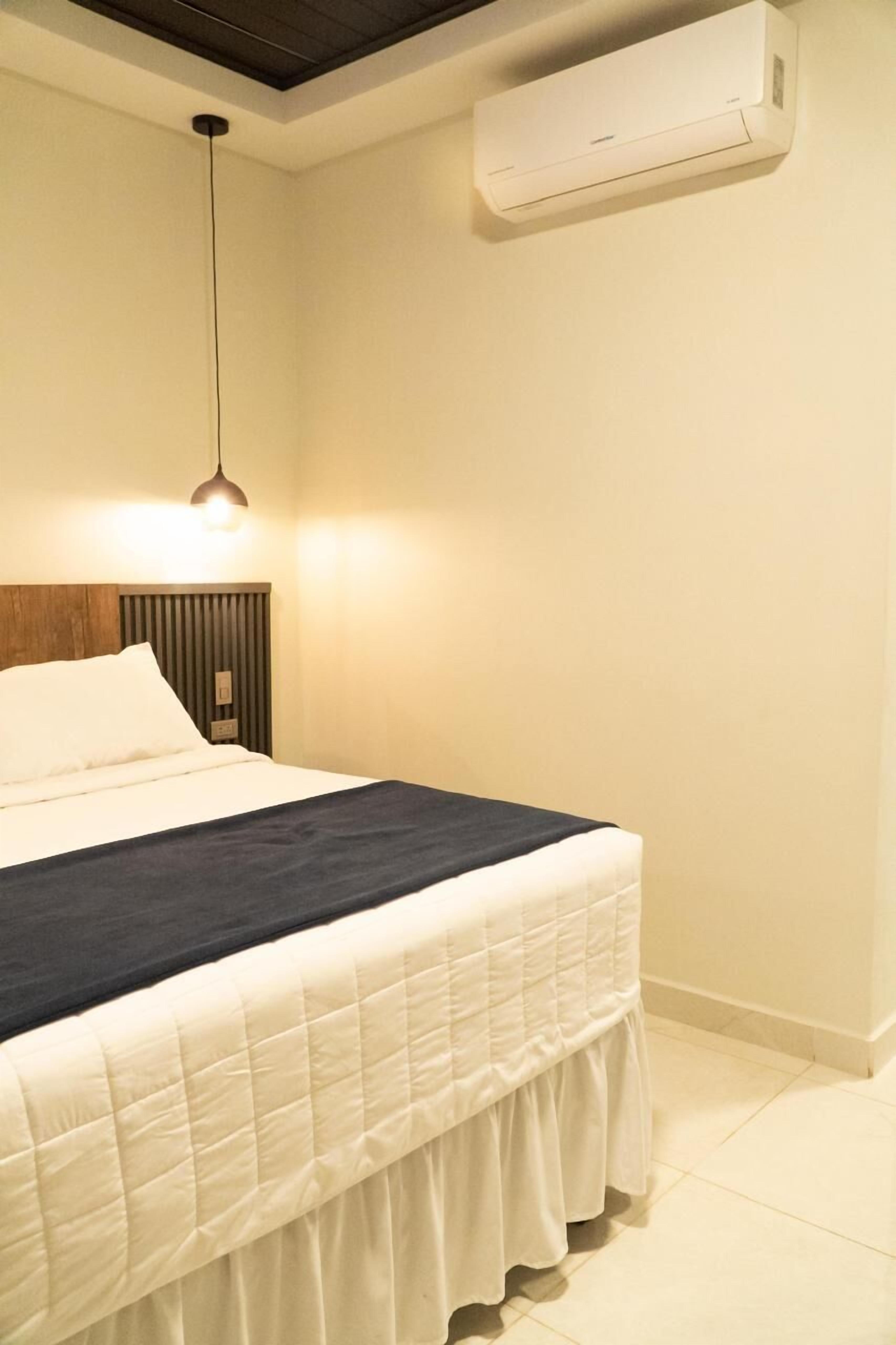 A bedroom with a bed, a hanging light fixture, a wall-mounted air conditioner, and a wooden headboard.