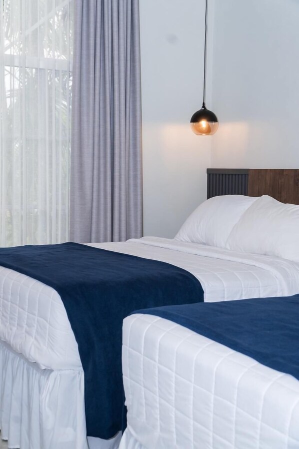 A hotel room with two beds, a window with sheer curtains, and a hanging pendant light.