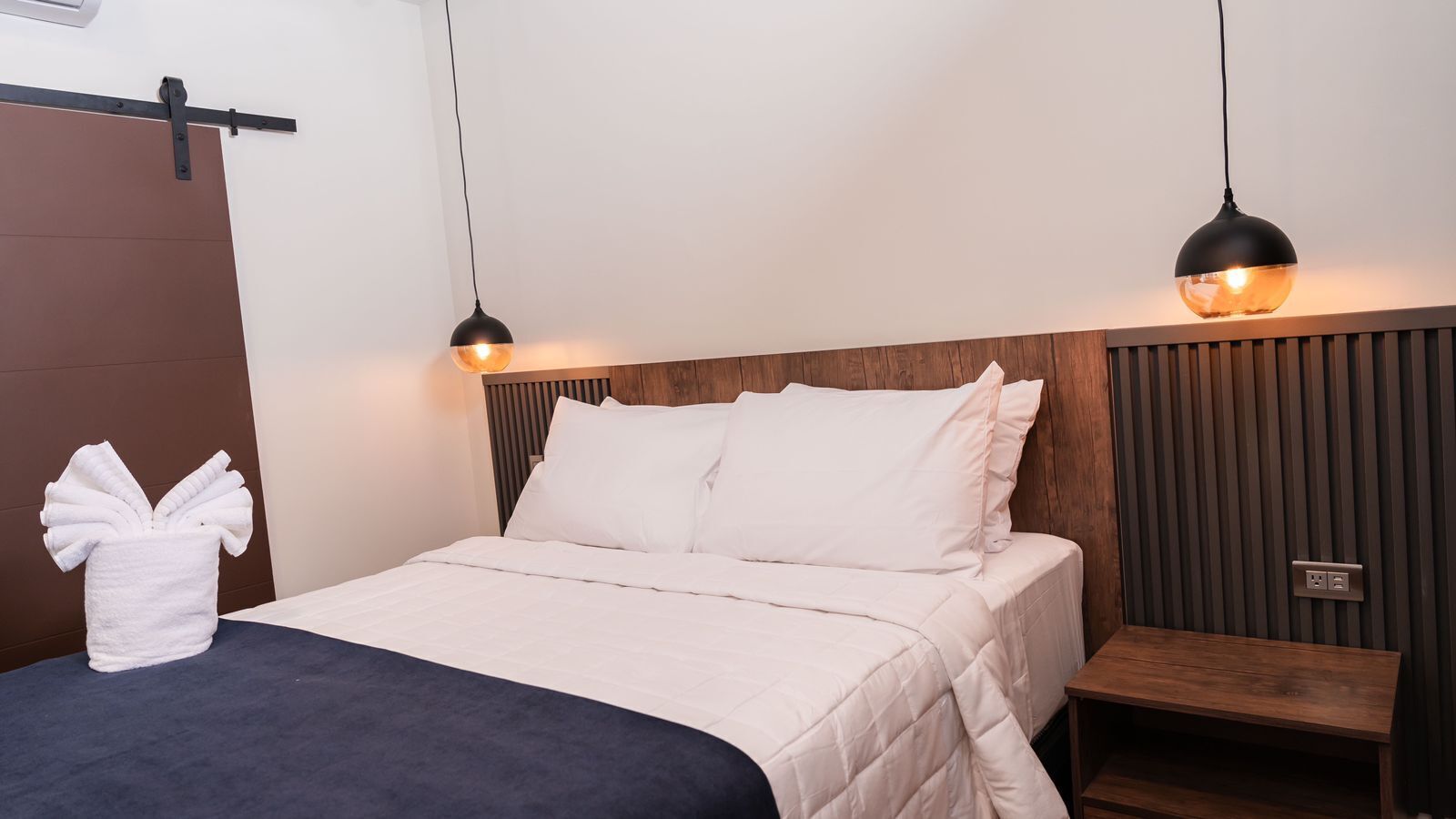A neatly made bed with white linens, a wooden headboard, and two pendant lights.