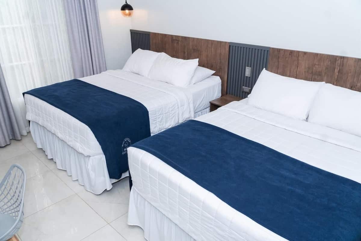 A hotel room with two beds, each with white bedding and a blue accent. There is a chair and a nightstand with a lamp.