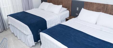 Habitación de hotel con dos camas, cada una con ropa blanca y un detalle azul. Hay una silla y una mesita de noche con una lámpara.