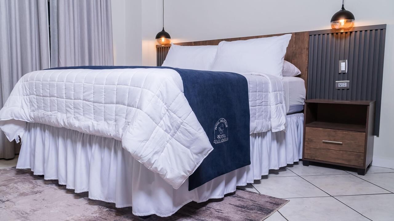 A neatly made bed with a white and blue bedspread, a wooden nightstand with a drawer, and a hanging pendant light.