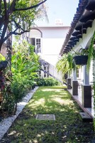 Un sendero estrecho bordeado por abundante vegetación y arquitectura tradicional, que conduce a un edificio con techo de tejas y unidad de aire acondicionado visible.