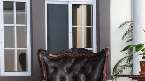 A dark brown, tufted leather armchair with ornate wooden carvings, positioned in front of a window with white frames and a dark wall. - Hotel Boutique Nueva Valladolid (Comayagua)