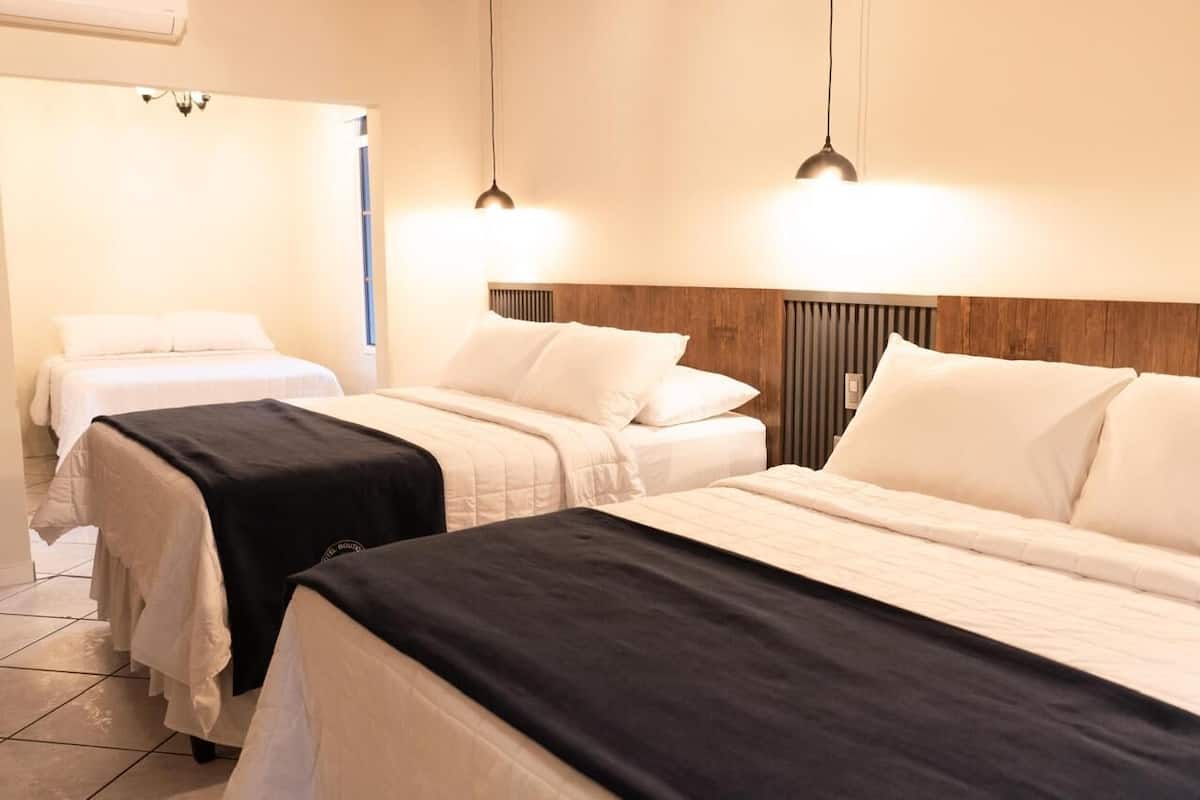 A hotel room with two beds, each with white bedding and a black headboard. There are pendant lights above the beds, and the room has a wooden headboard on one side.