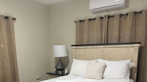 Una cama bien tendida con sábanas blancas, una lámpara de noche y un aire acondicionado de pared.