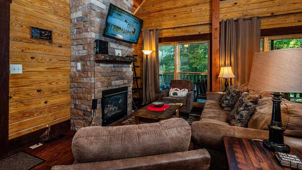 A cozy cabin interior with a stone fireplace, wooden walls, a comfortable sofa, and a television mounted on the wall. - 3 Irish Sons, 4BR, 3 BA  Log Cabin in the woods. (Nashville)