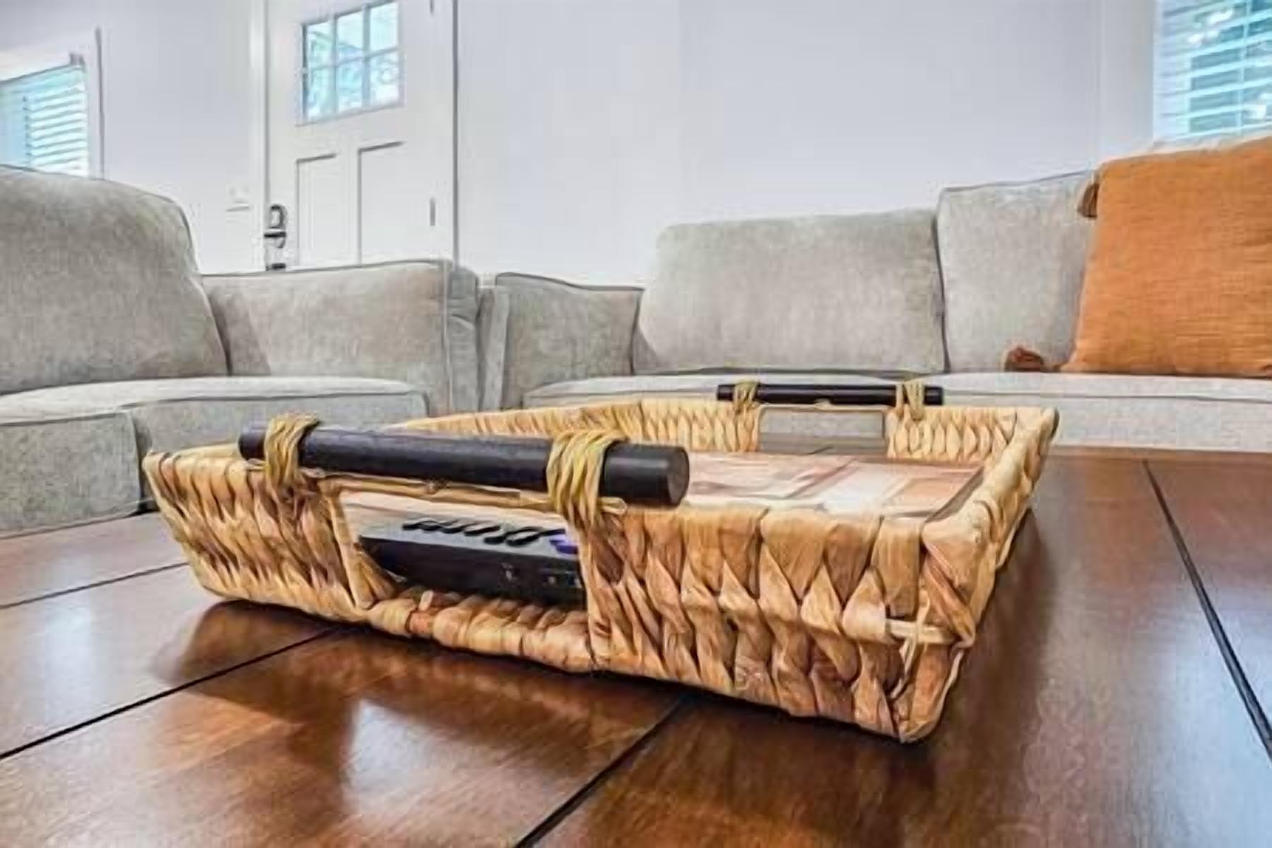 Ein geflochtener Korb mit Fernbedienung auf einem Holzboden in einem Wohnzimmer mit grauem Sofa und einem Fenster im Hintergrund.