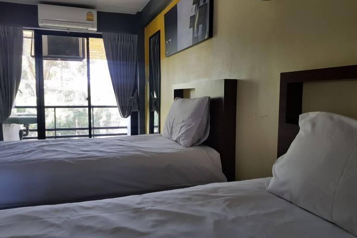 A hotel room with two beds, a window with curtains, an air conditioning unit, and a wall-mounted artwork.