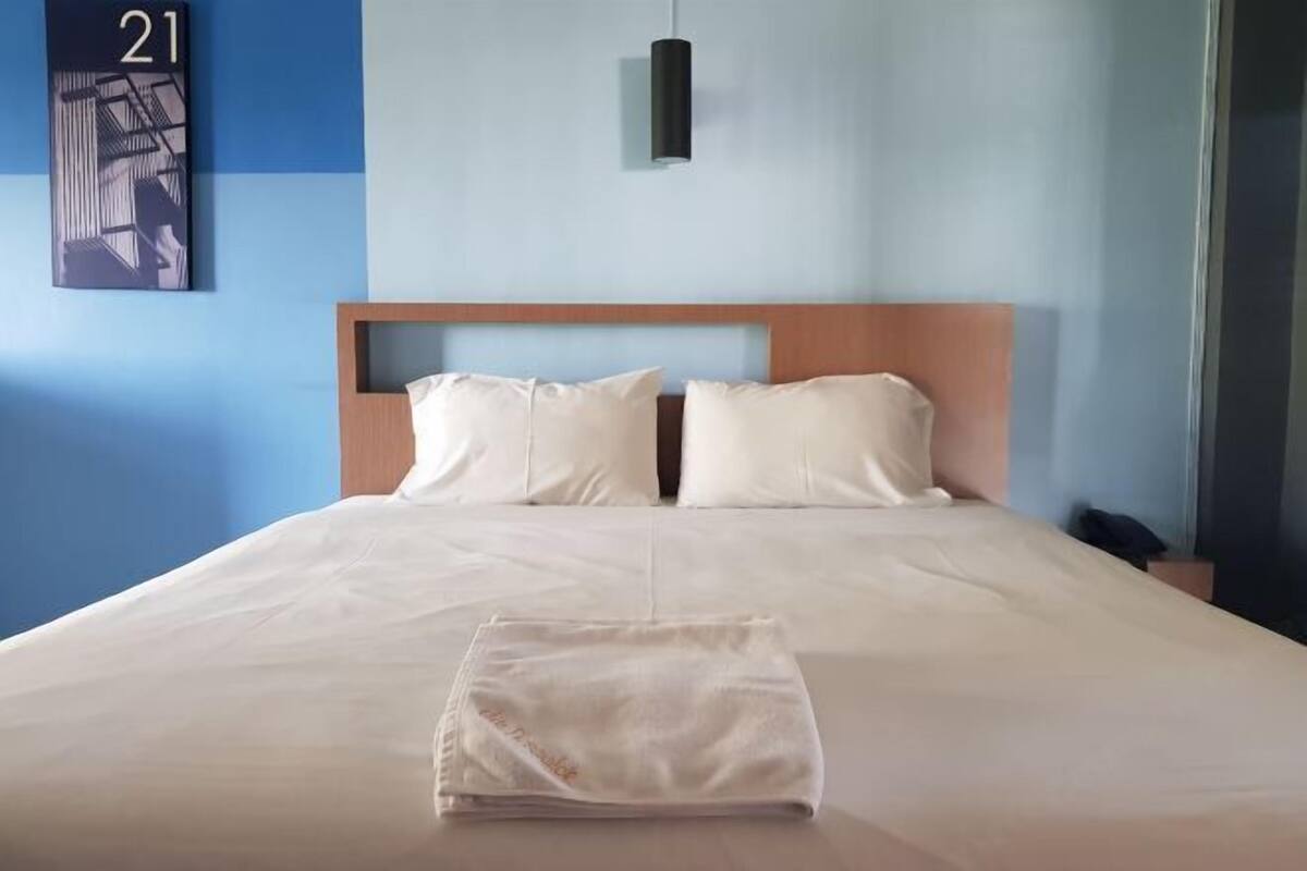 A hotel room with a large bed, a headboard, a wall-mounted light, and a number on the wall.