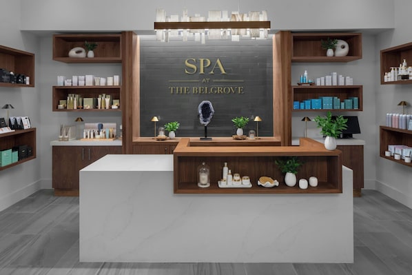 Zona de recepción de spa con mostrador central, estanterías de madera con productos de spa y un cartel que dice "SPA AT THE BELGROVE".