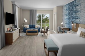 Een moderne hotelkamer met een groot bed, een bank, een bureau en een balkon met uitzicht op palmbomen.