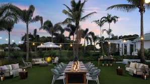 An outdoor dining area with a long table set for six, surrounded by white wicker chairs, under string lights. - The Belgrove Resort & Spa, West Palm Beach, Autograph Collection (West Palm Beach)