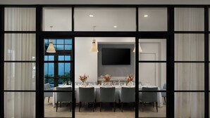 A dining area with a large table set for six, a flat-screen TV mounted on the wall, and a view of the outdoors through large windows. - The Belgrove Resort & Spa, West Palm Beach, Autograph Collection (West Palm Beach)