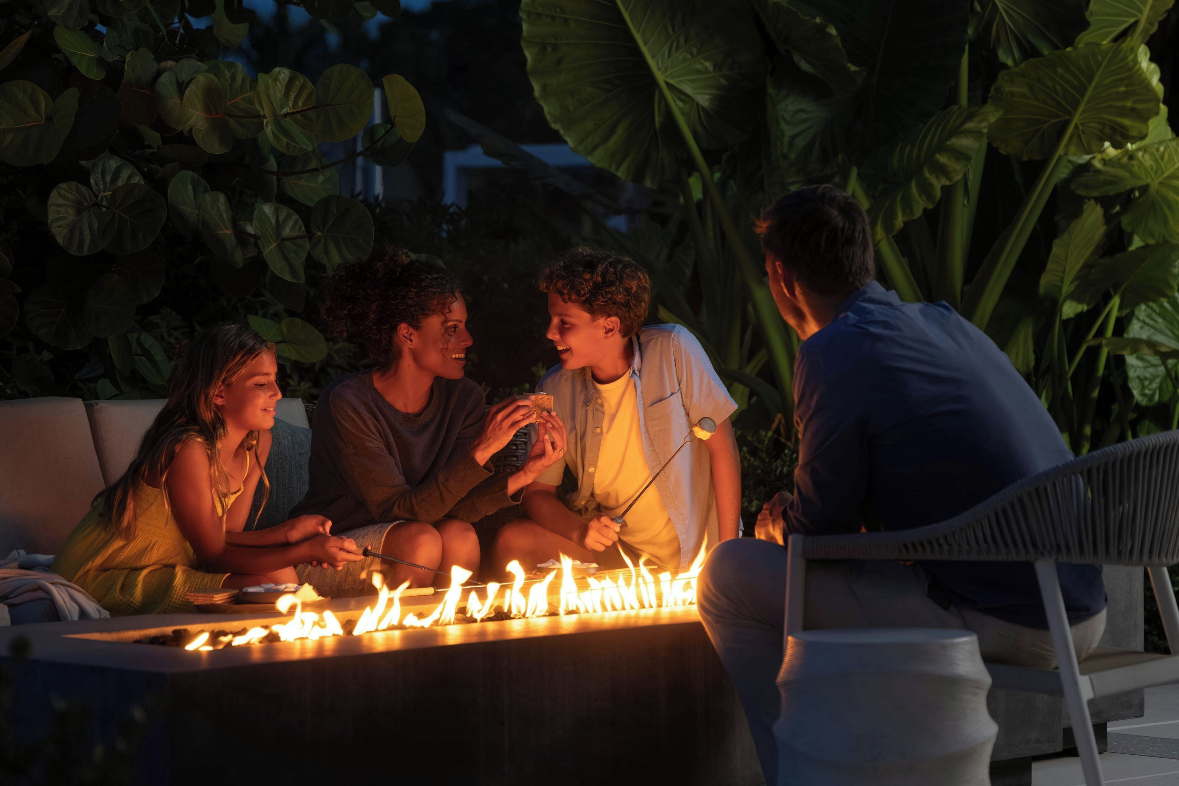A group of people sitting around a fire pit outdoors with large green leaves in the background.