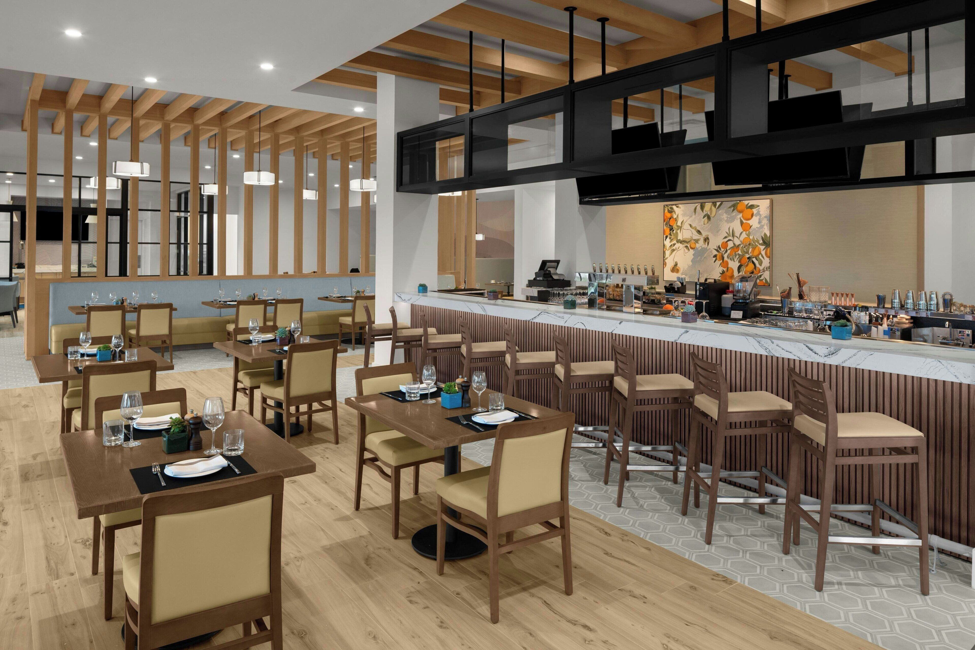 A modern restaurant with wooden tables and chairs, a bar area, and a large window providing natural light.