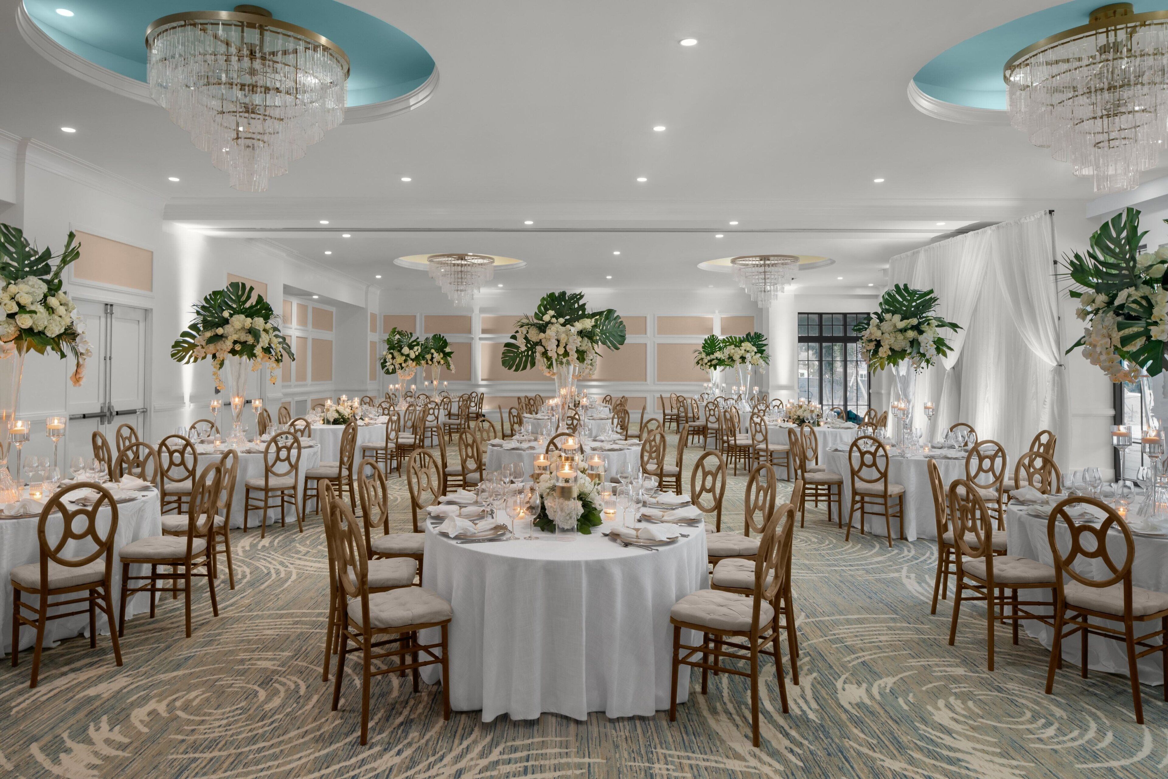 Un salón de banquetes con mesas redondas, manteles blancos y centros de mesa florales.