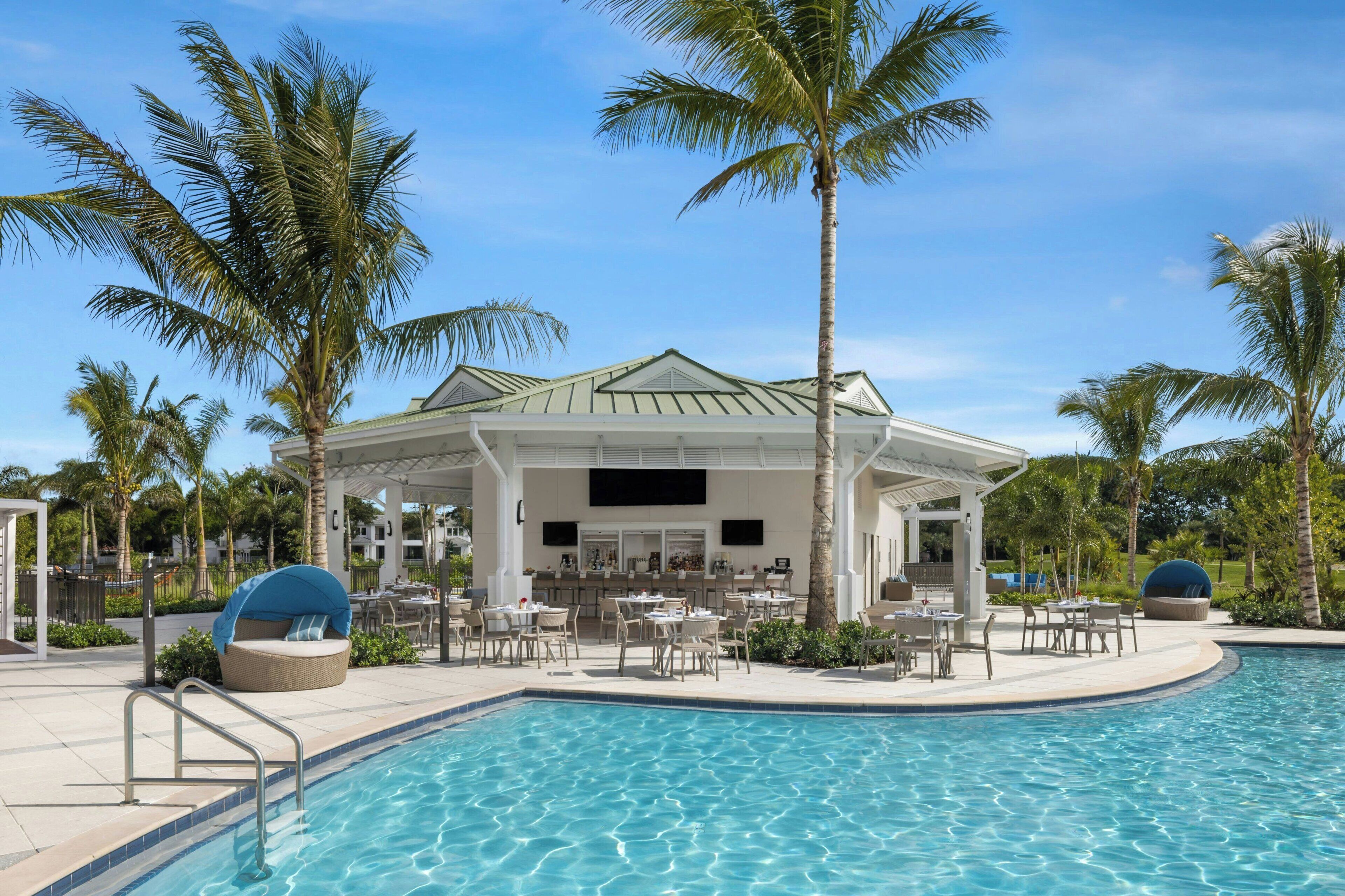 A poolside area with a covered seating area, palm trees, and a bar.