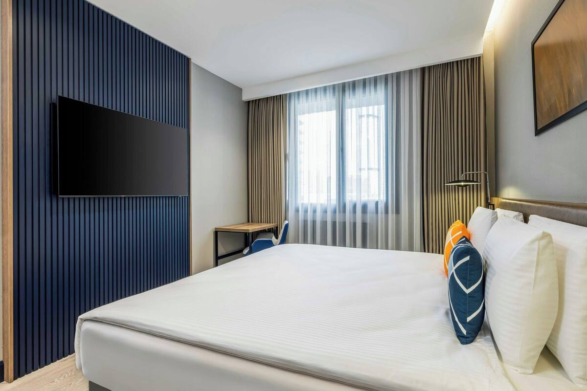 A hotel room with a large bed, a flat-screen TV mounted on the wall, a desk with a chair, and a window with curtains.