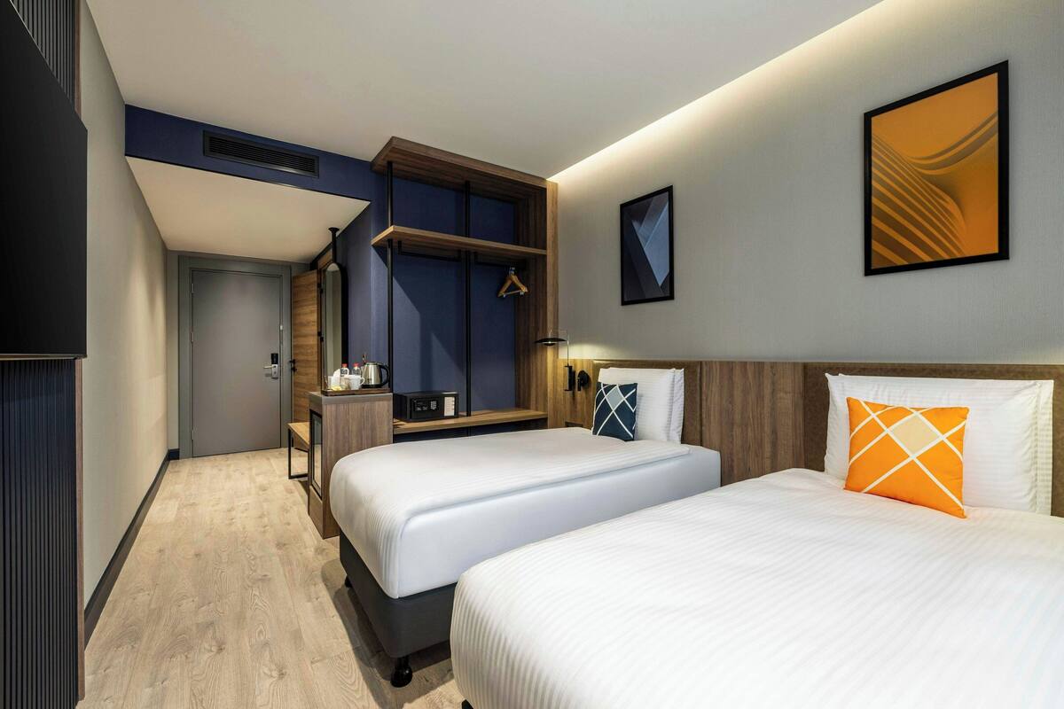 A hotel room with two beds, a wooden headboard, a bedside table with a lamp, and a wall-mounted artwork.