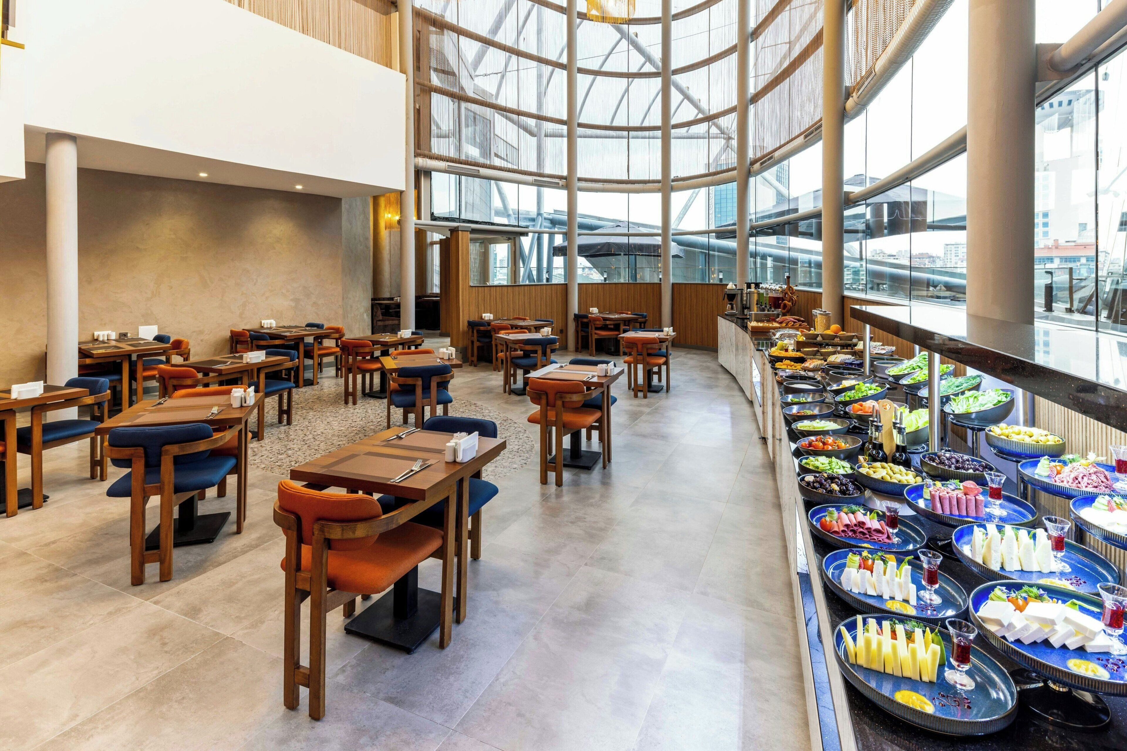 Un vaste espace intérieur aménagé en salle à manger, avec de nombreuses tables et chaises, un buffet proposant une variété de plats et de grandes fenêtres offrant une vue sur la ville.