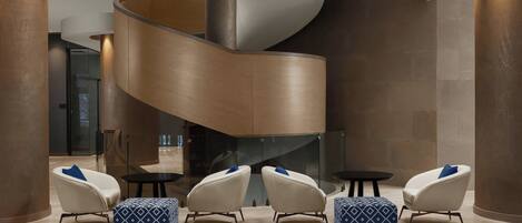 A modern lobby with a curved staircase, seating area with patterned ottomans, and a glass wall.