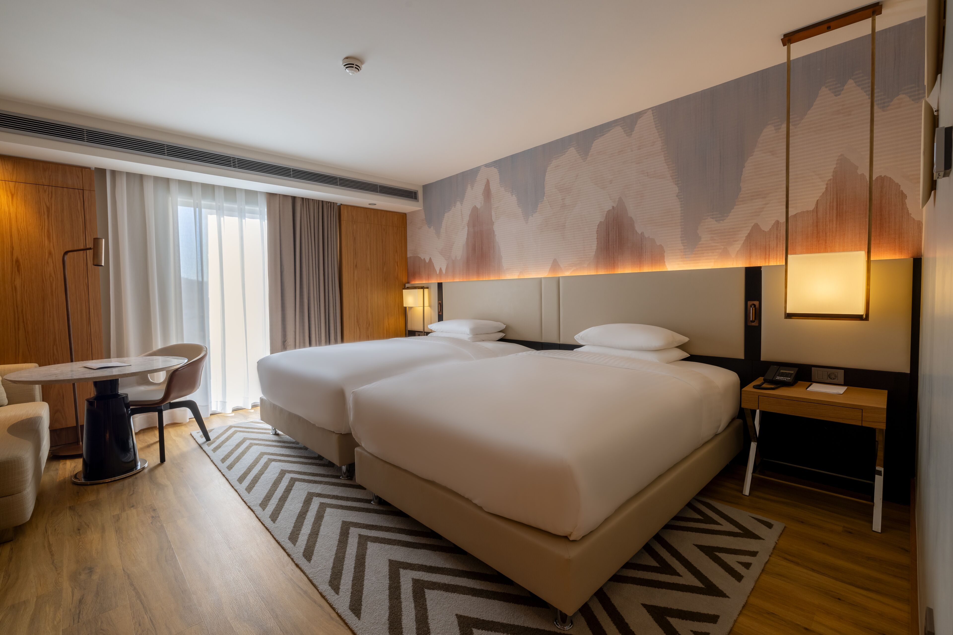 A hotel room with two beds, a desk, a chair, and a large wall mural.
