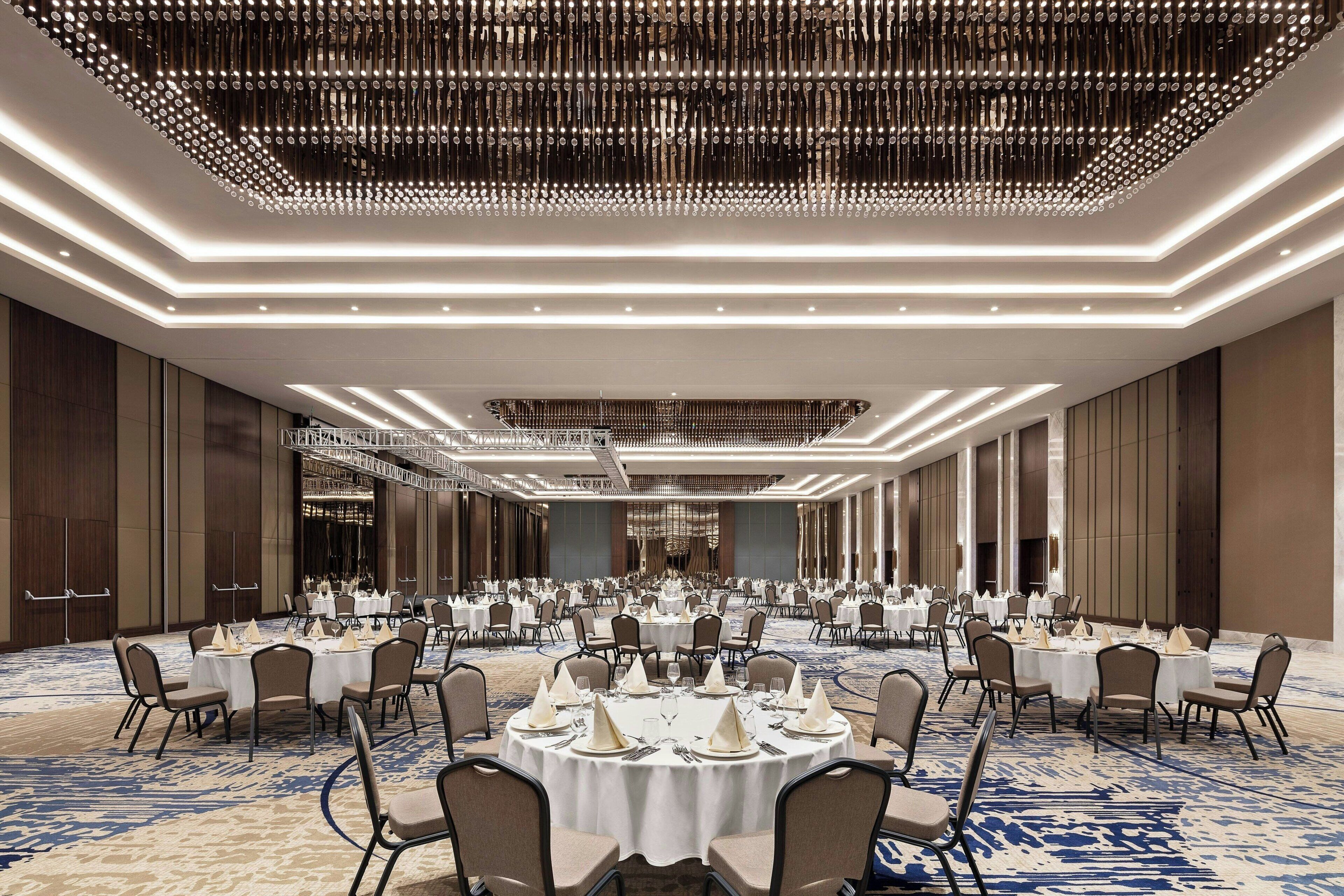 A large banquet hall with round tables set for dining, a modern chandelier, and a carpeted floor.