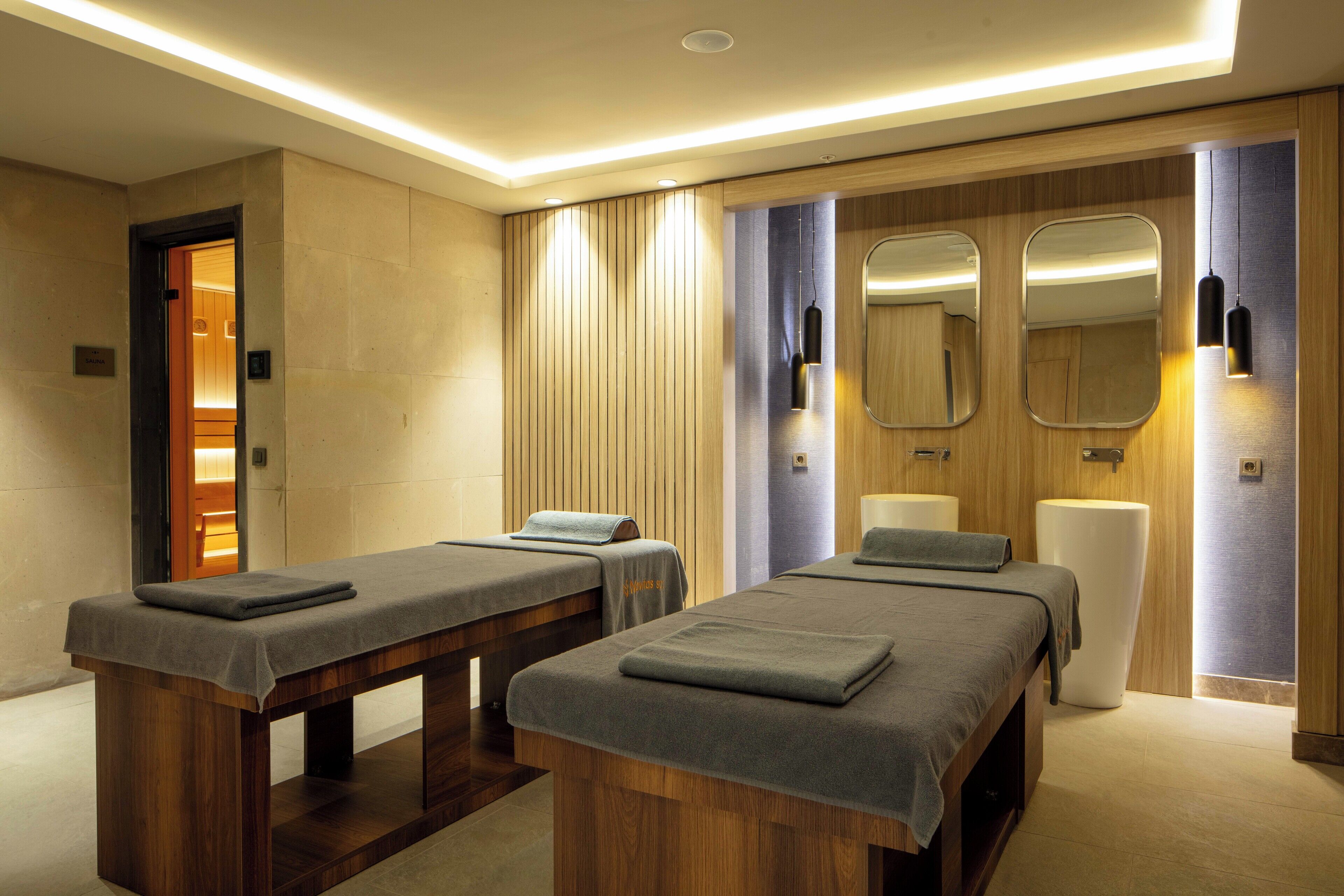 A modern spa room with two massage tables, wooden paneling, and a large mirror.