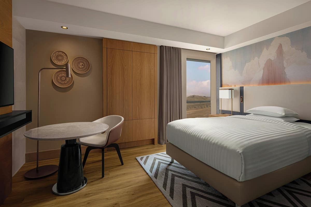 A modern hotel room with a bed, a desk, and a chair. There is a large window with a view of the mountains.
