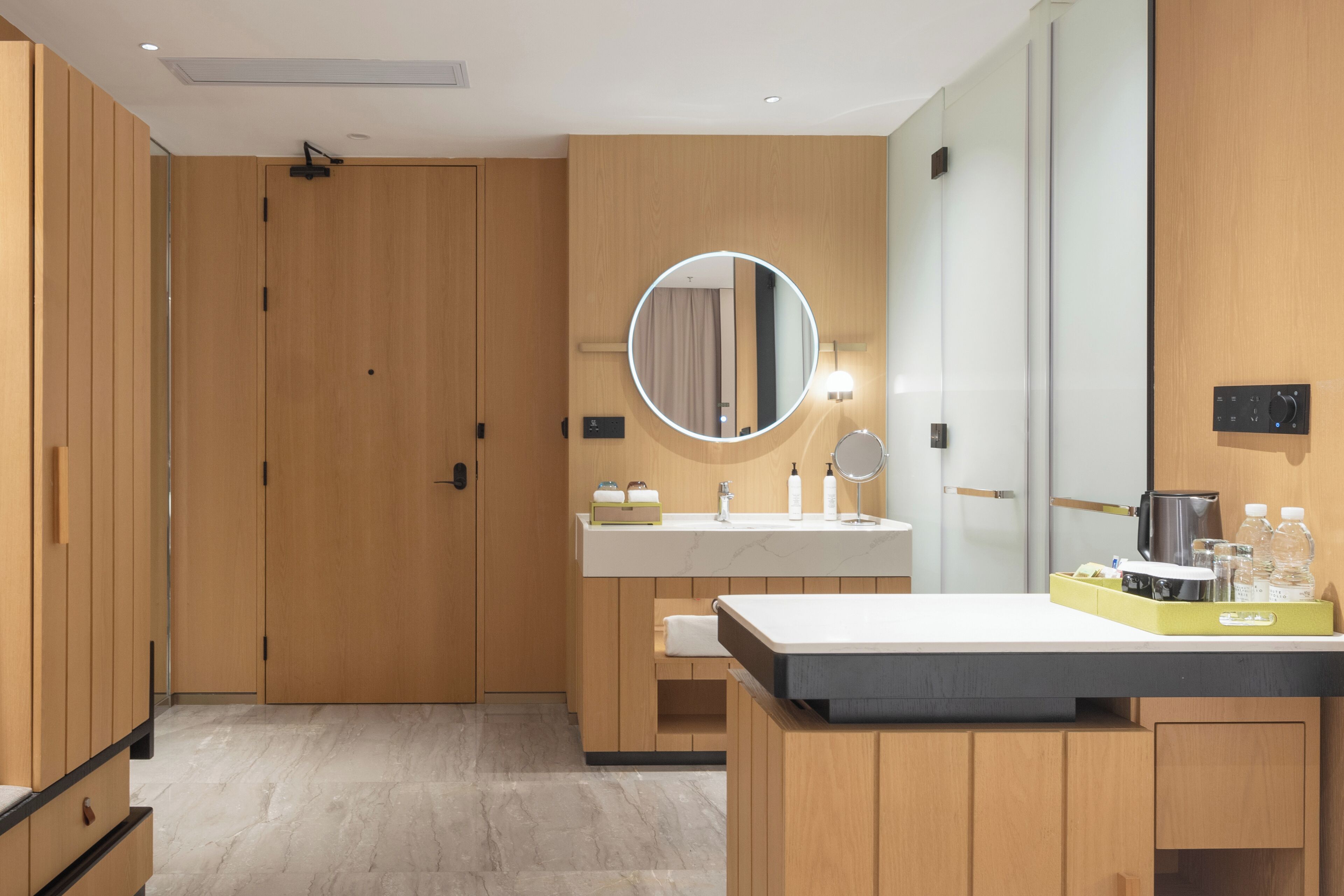 A modern bathroom with wooden cabinetry, a round mirror, a sink with toiletries, and a coffee maker.