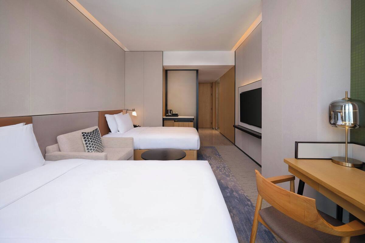 A hotel room with two beds, a desk, a chair, a TV, and a small kitchenette.