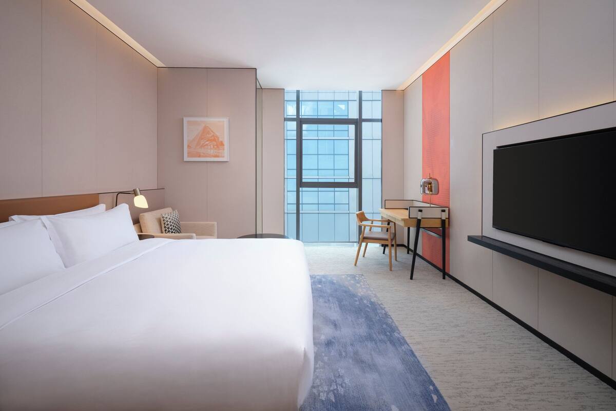 A hotel room with a large bed, a desk, a chair, a TV, and a window with a view.