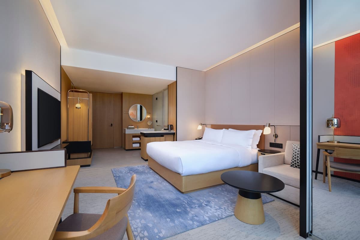 A modern hotel room with a large bed, a desk, a chair, a TV, and a bathroom area.