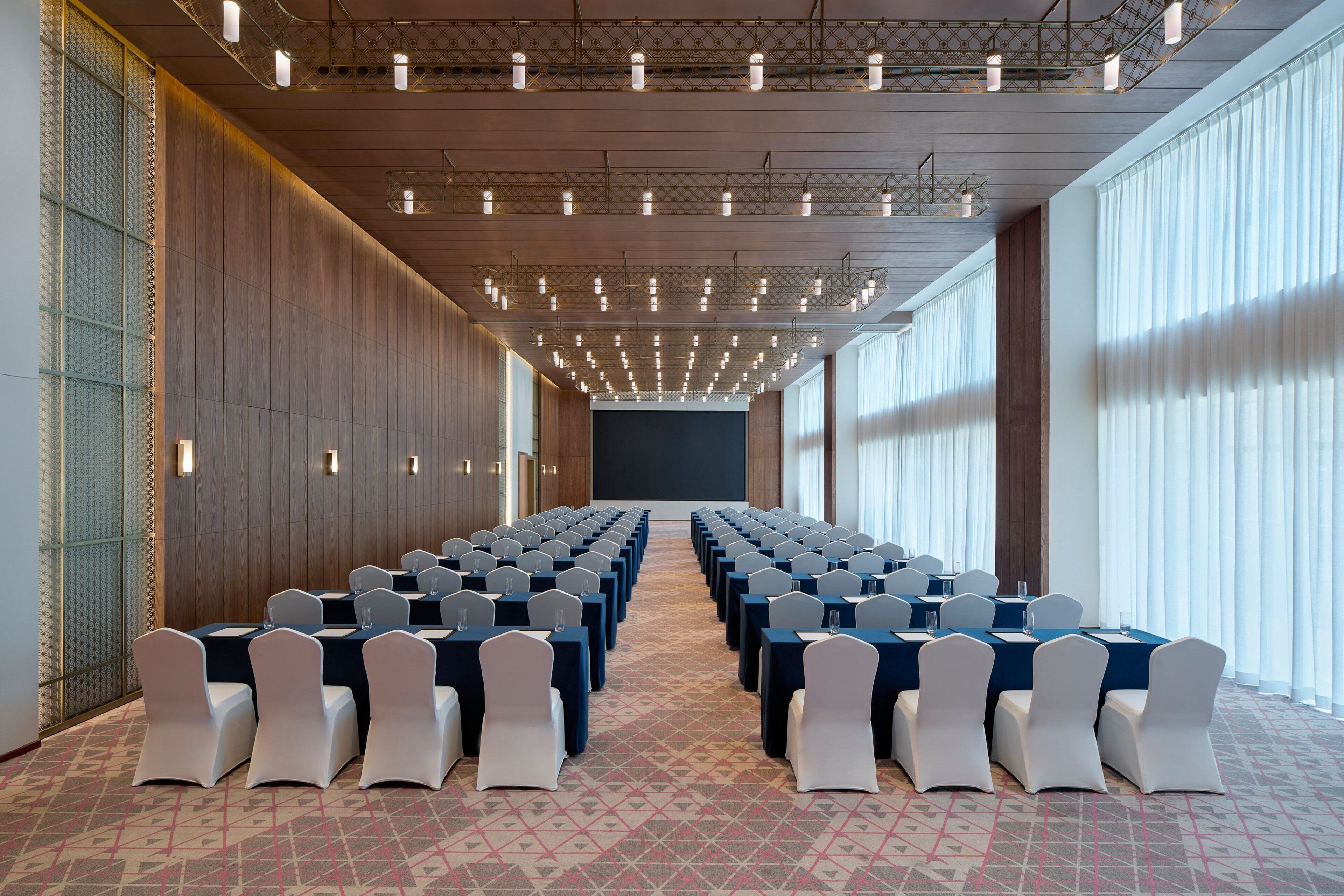 Une salle de conférence avec des rangées de tables et de chaises, un écran de projection et une moquette à motifs.
