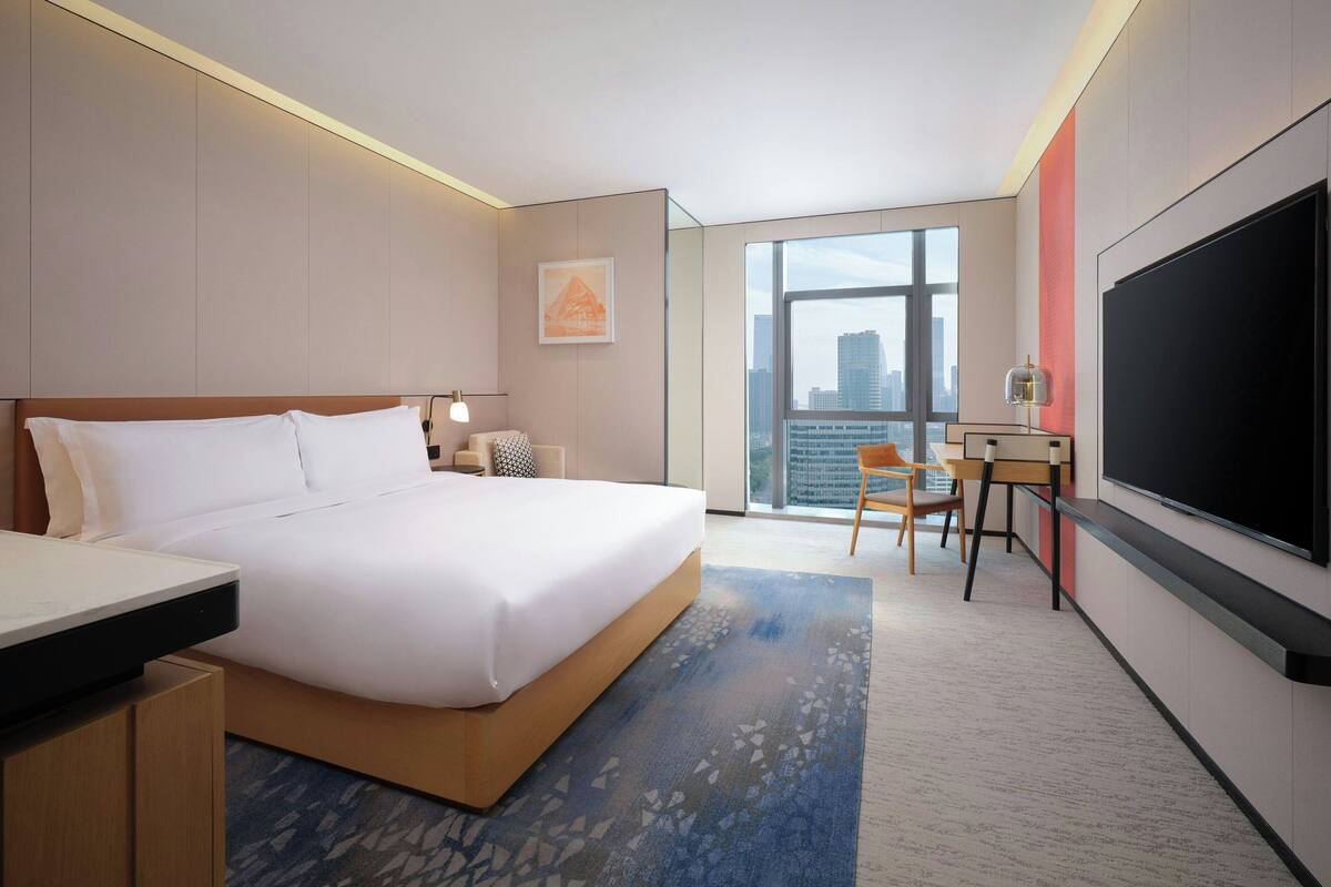A modern hotel room with a large bed, a desk, a flat-screen TV, and a city view.