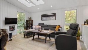 Un salon spacieux avec un canapé gris, une table basse en bois et un fauteuil noir. Un téléviseur à écran plat est fixé au mur, et une grande fenêtre offre une vue sur la verdure à l’extérieur.