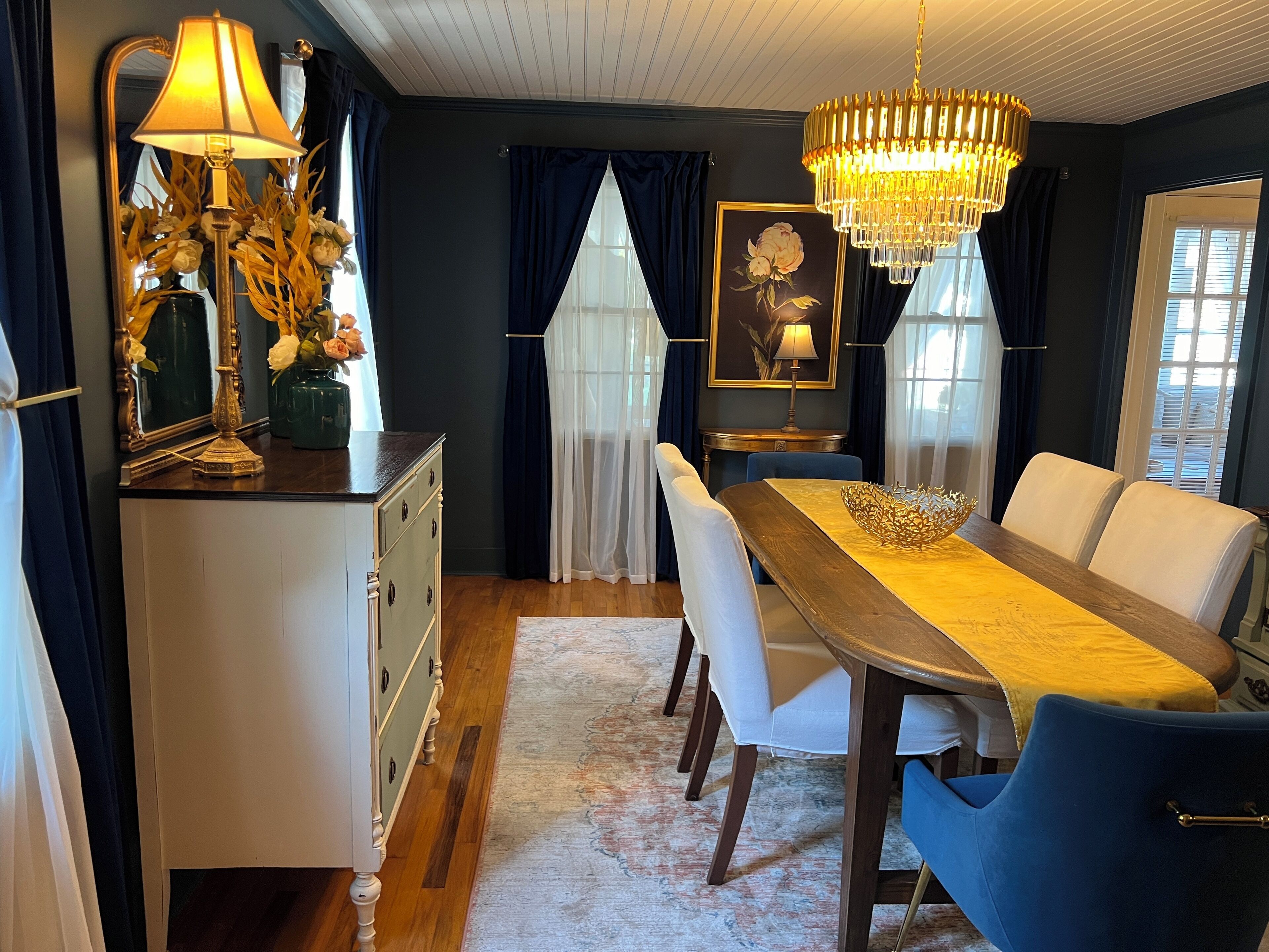 A dining area with a wooden table, white chairs, a chandelier, and a mirror with flowers.