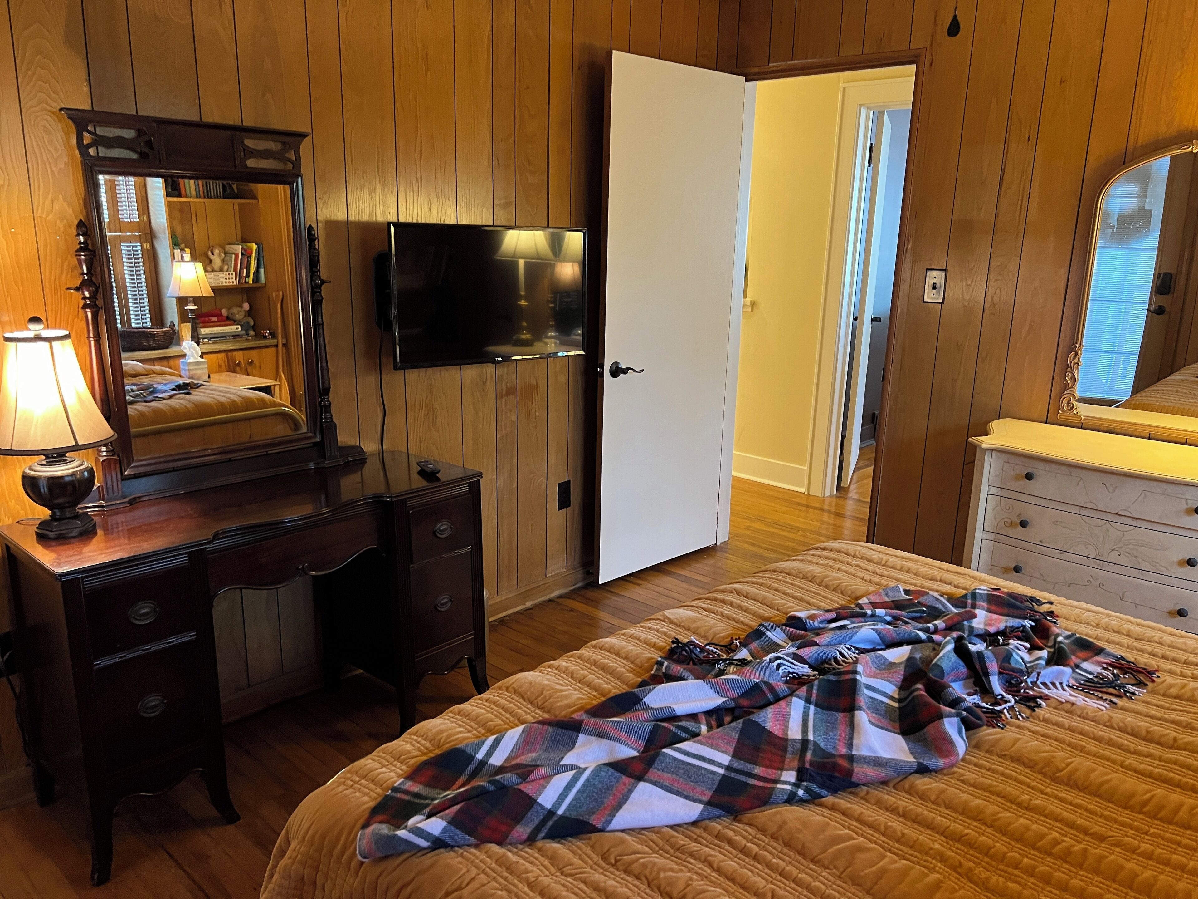A bedroom with wooden paneling, a bed with a plaid blanket, a dresser, a desk with a lamp, a TV, and a mirror.