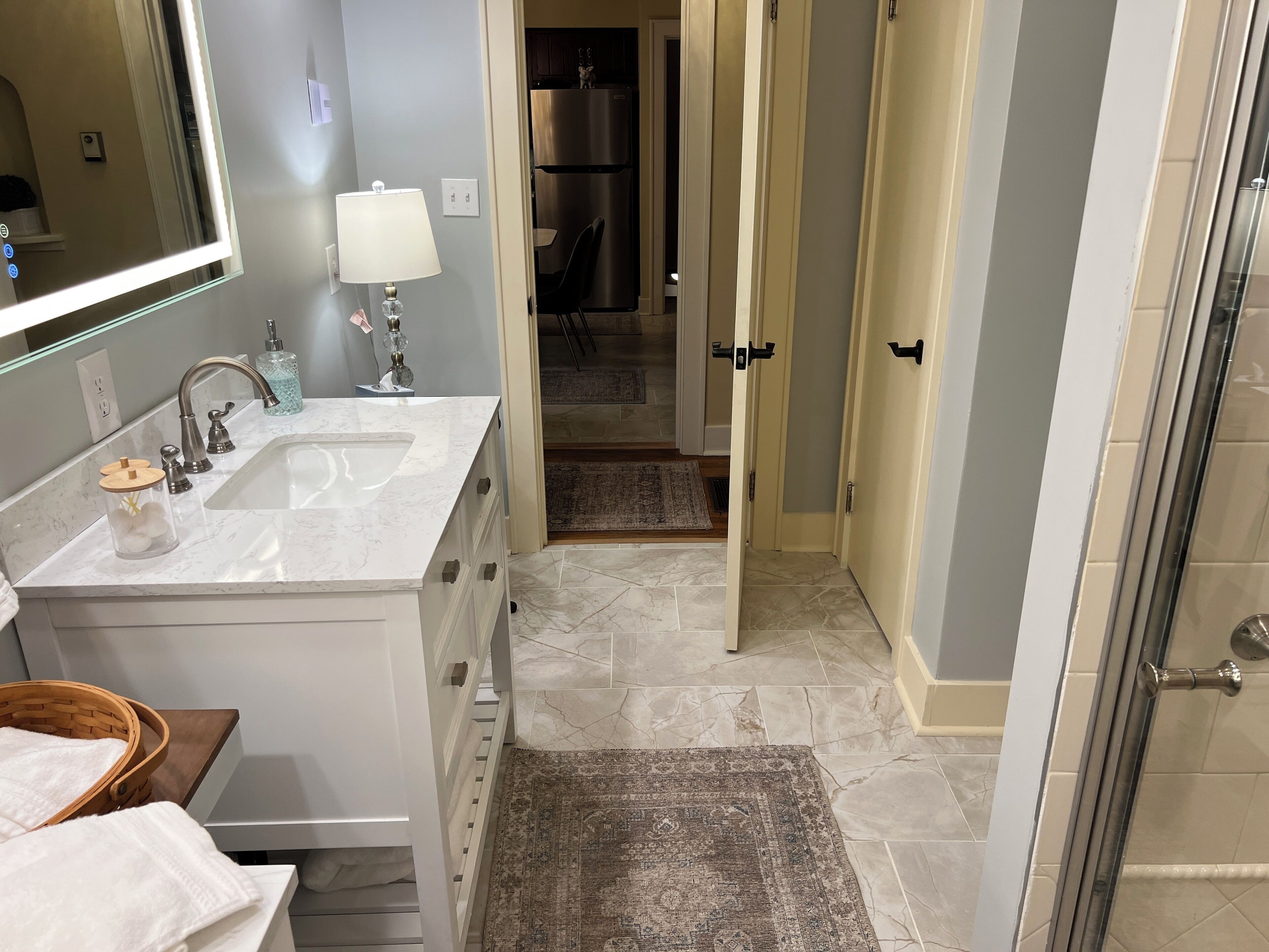 A modern bathroom with a large mirror, a sink, and a vanity. There is a rug on the floor and a basket with towels.