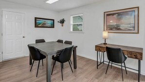 Een kamer met een eettafel, stoelen, een bureau met stoel, een lamp, een deur, een raam en twee ingelijste foto's aan de wanden.