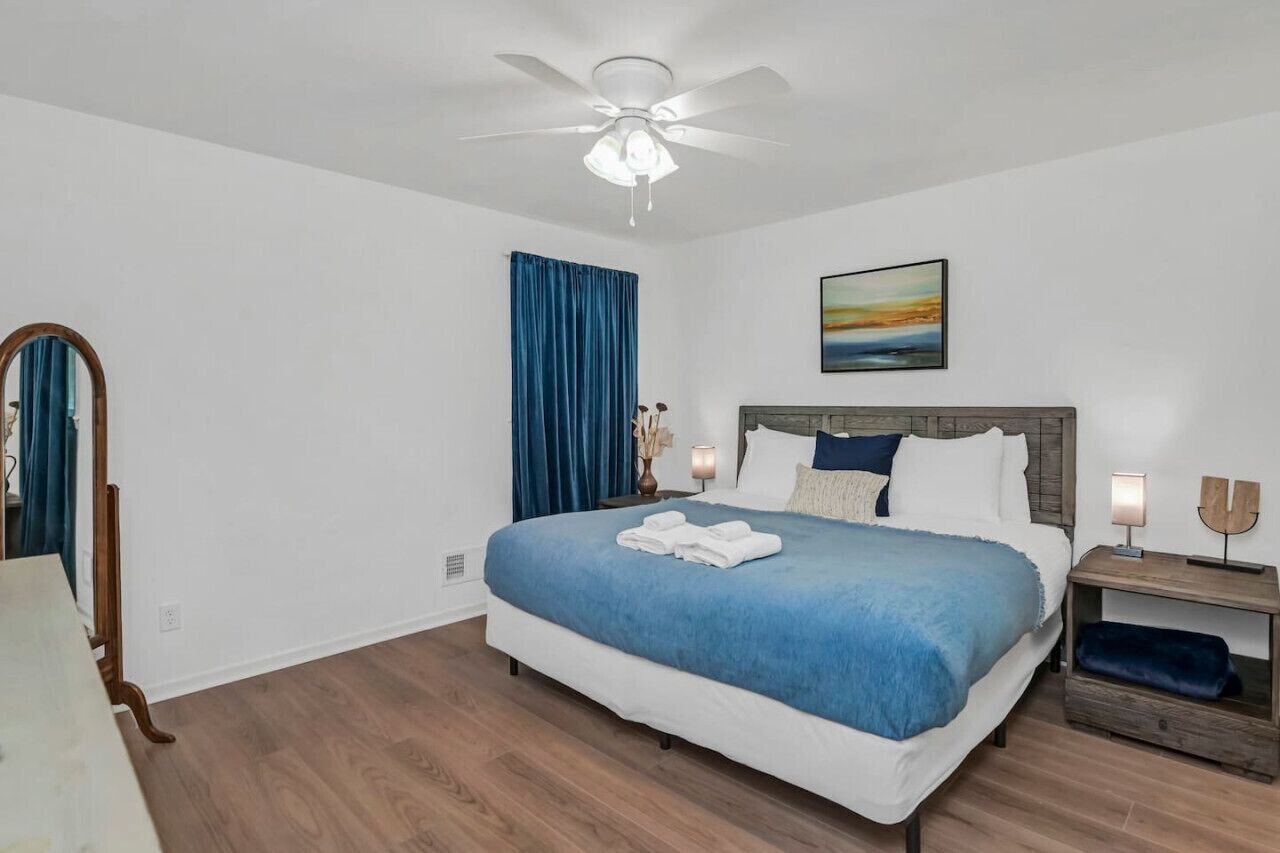 Un dormitorio con cama, ventilador de techo, mesita de noche con lámpara, un cuadro en la pared y una ventana con cortinas azules.
